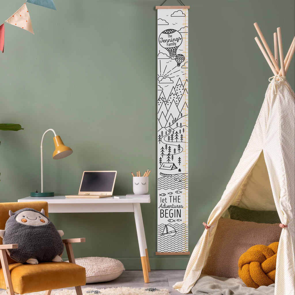 A canvas height chart with a mountain and hot air balloon design, personalized with the text 'The Smith Family'. It is displayed in a room setting with a wooden bar at the top and bottom for hanging, and a leatherette string is attached for wall mounting.